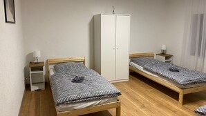 A room with two single beds, a wardrobe, a small table with a lamp, and a nightstand with a lamp.