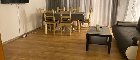 A living room with a wooden floor, a dining table with chairs, a sofa, and a TV mounted on the wall.