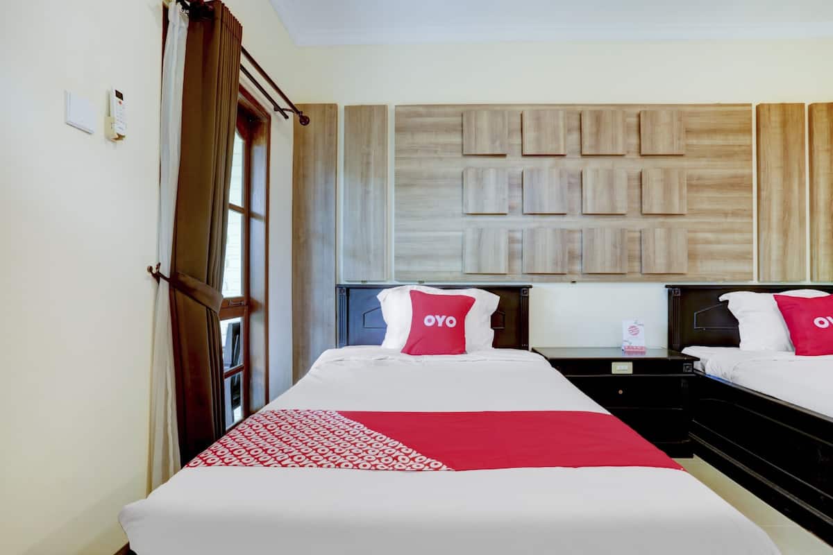 A hotel room with two beds, each with a red pillow labeled "OYO," a patterned red and white bedspread, and a wooden panel wall design.