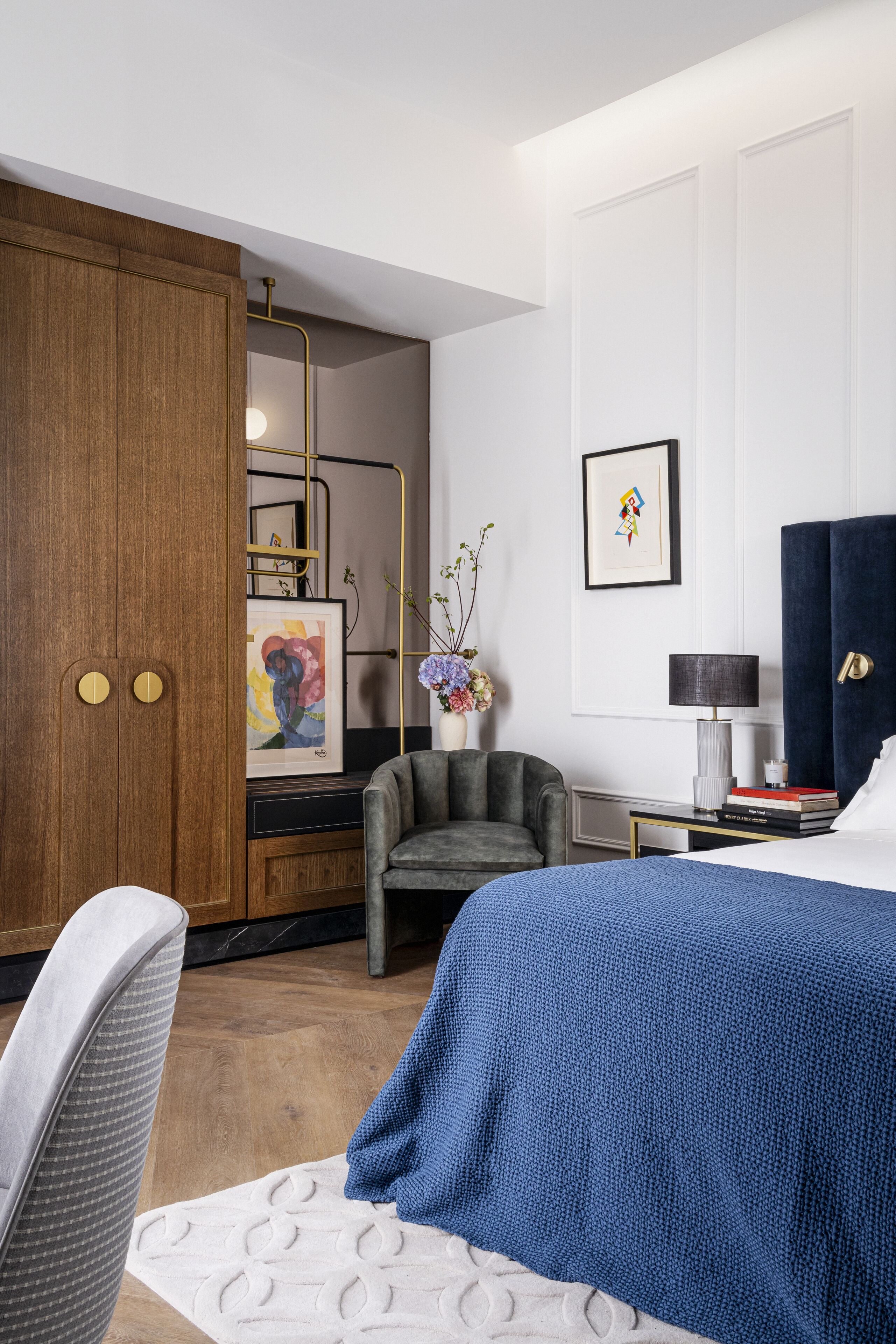 A hotel room with a large wardrobe, a grey armchair, a bed with a blue blanket, a small table with a lamp, and a framed artwork on the wall.