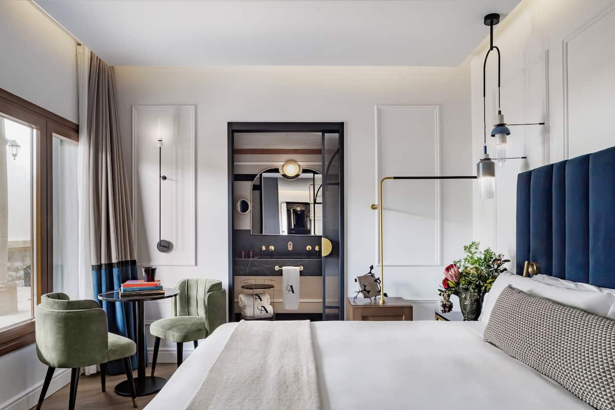 Una habitación de hotel moderna con una cama grande, dos sillones verdes, una mesita con libros, un tocador con espejo y vistas al exterior a través de una ventana.