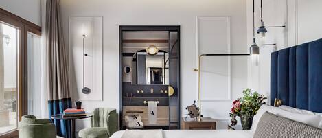 A modern hotel room with a large bed, two green armchairs, a small table with books, a vanity with a mirror, and a view of the outdoors through a window.