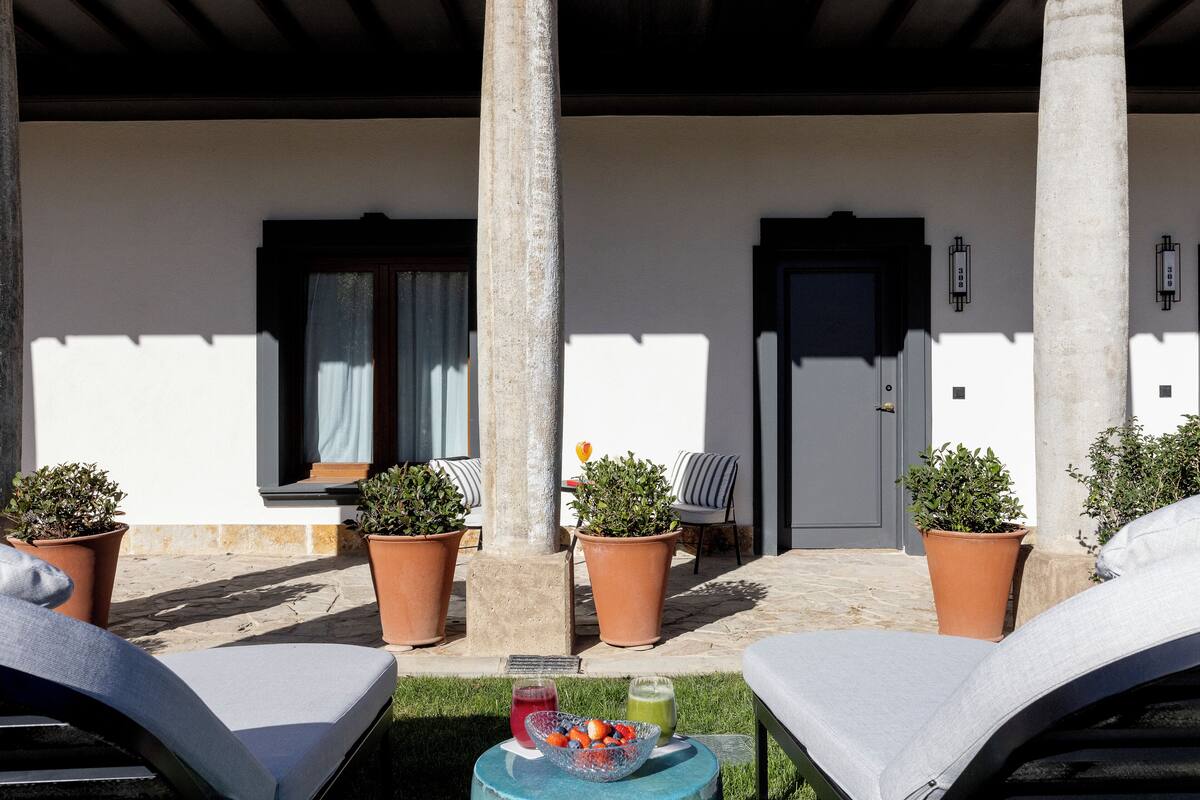 Un espacio de estar al aire libre con techo, sillones, plantas en macetas y una mesa pequeña con bebidas.