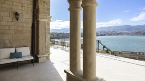 A stone balcony with columns, a bench, and a view of a city and water. - Palacio Arriluce Hotel (Getxo)