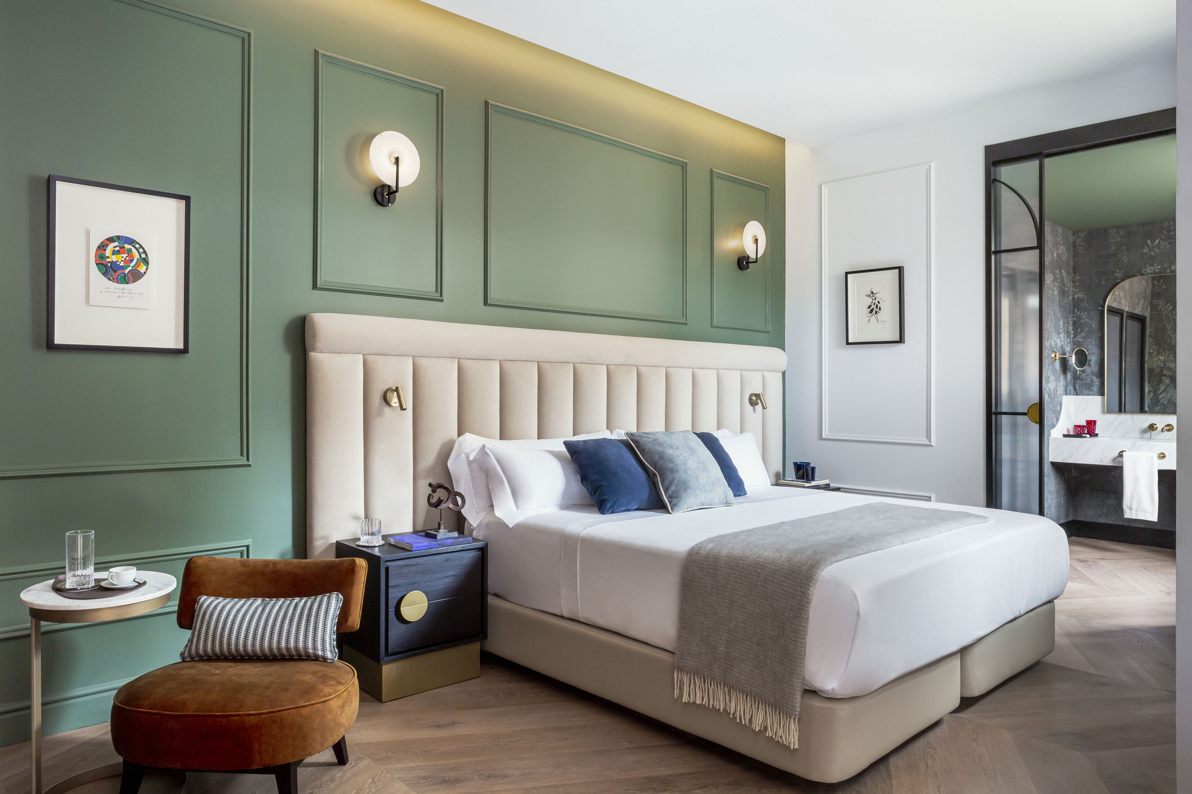 Un dormitorio moderno con una cama grande, una mesita de noche, una silla y un baño visible a través de una puerta abierta.