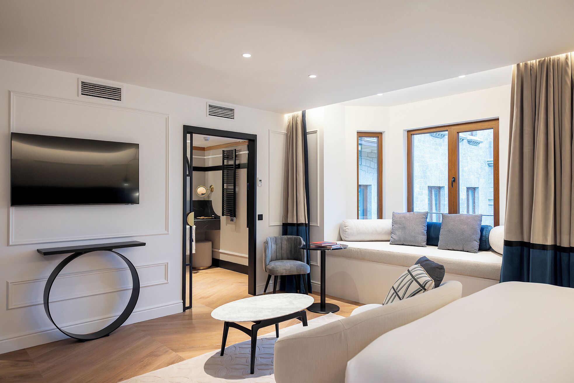 A modern hotel room with a flat-screen TV, a round table, a sofa with cushions, a chair, and a window with curtains.