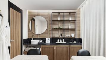 A modern hotel room with a wooden vanity, a round mirror, a black marble countertop, and two black chairs with towels.