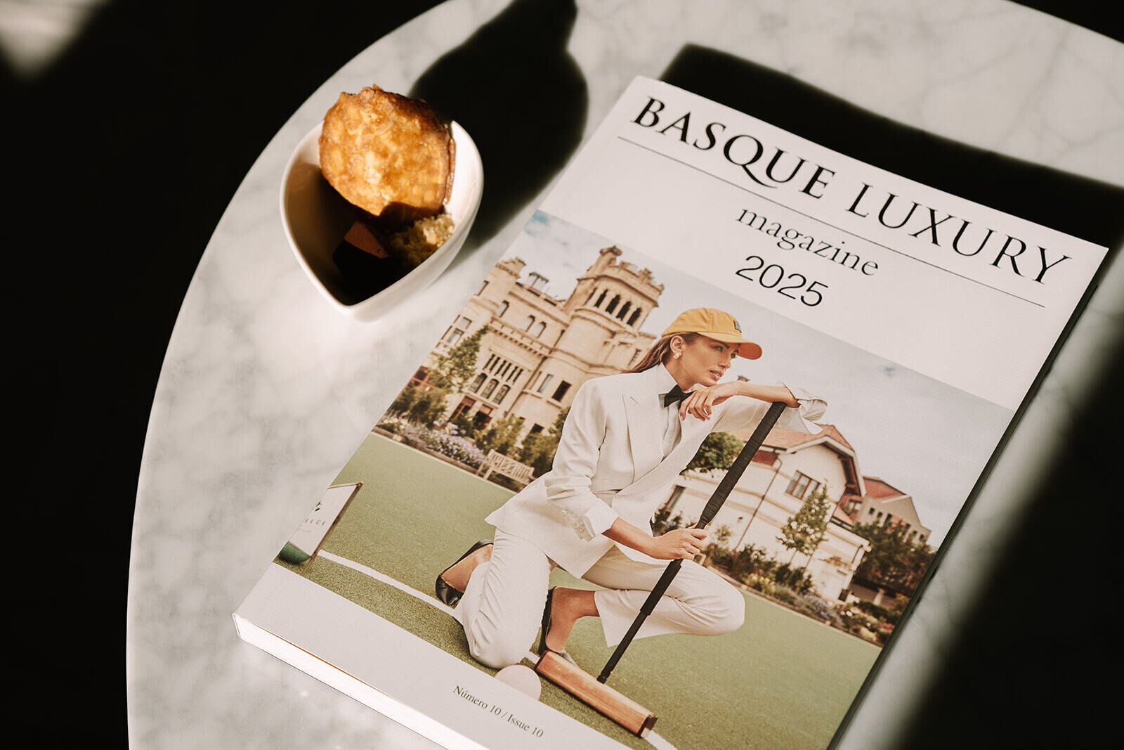 Una revista titulada 'BASQUE LUXURY' con una foto de portada de una persona con traje blanco sosteniendo un palo de golf, con un edificio y follaje de fondo.