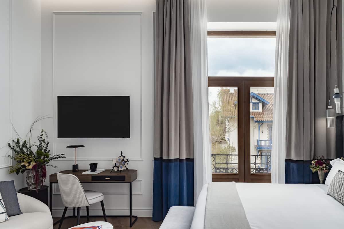 Una habitación de hotel con cama, escritorio con silla, televisor montado en la pared y ventana con cortinas.
