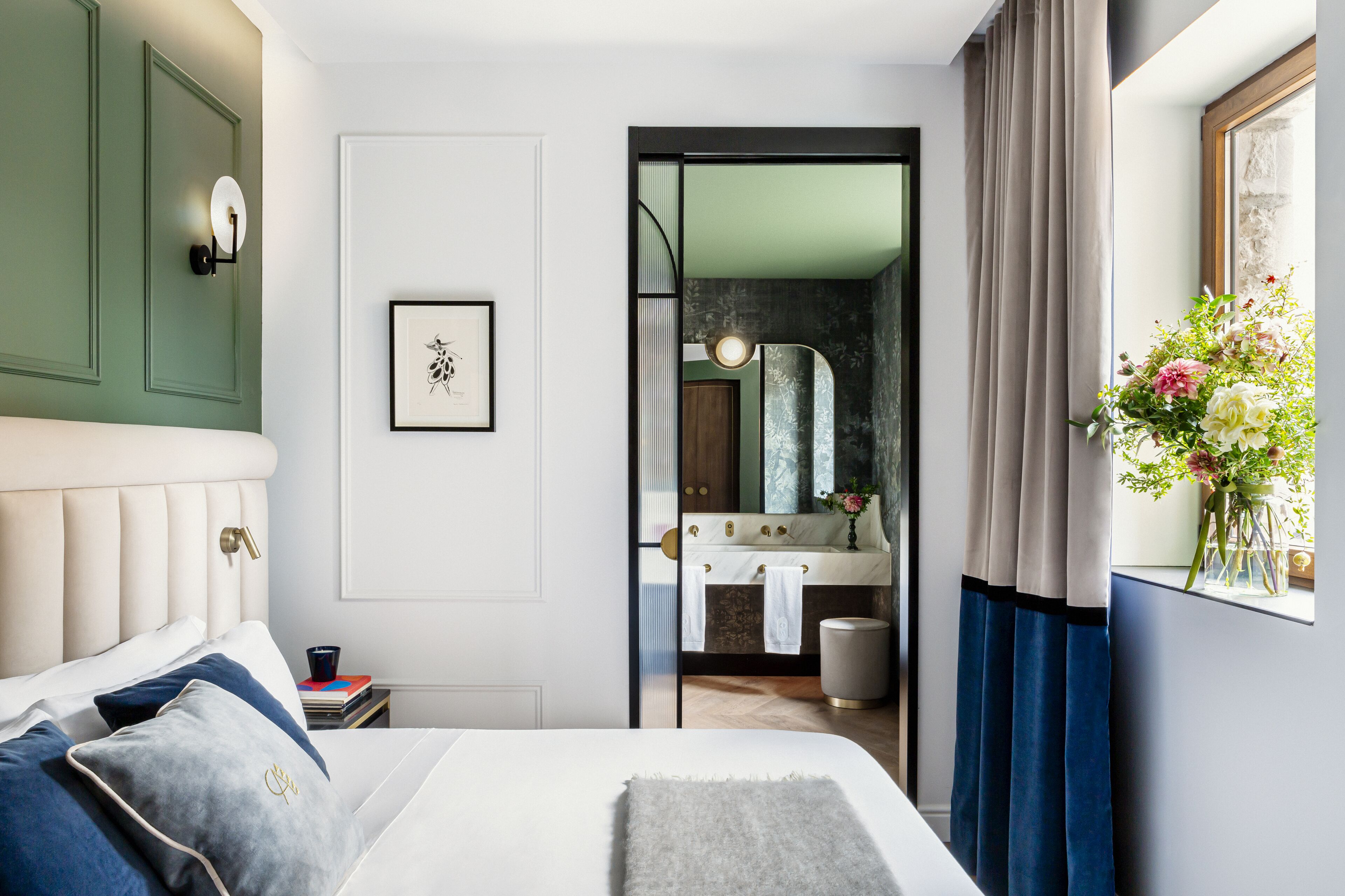 A bedroom with a bed, a bedside table, a nightstand, a mirror, a window with curtains, and a bathroom visible through a glass door.