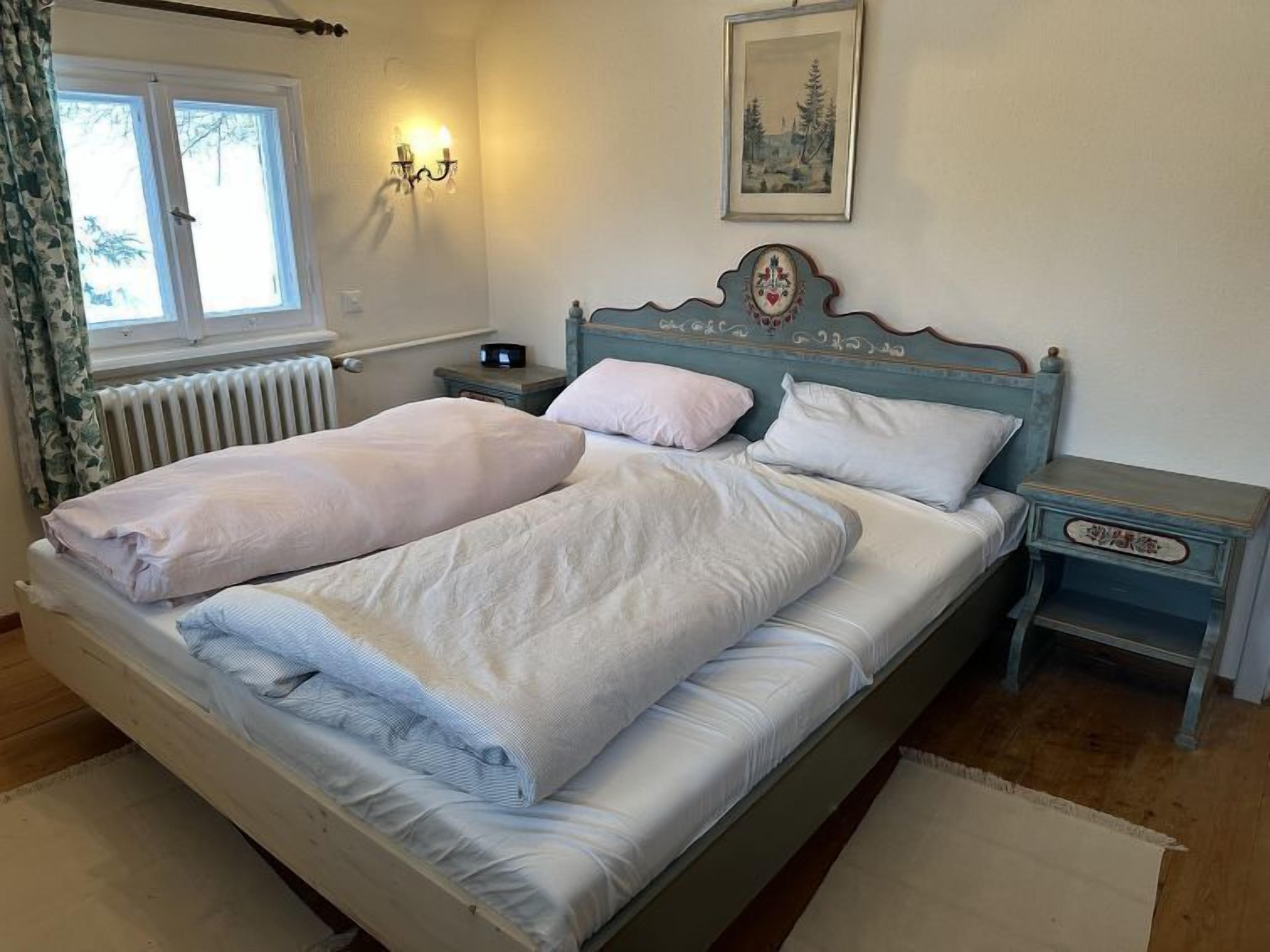 Un lit double avec du linge de lit blanc, une table de chevet avec une lampe, un tableau encadré au mur et une fenêtre avec des rideaux.