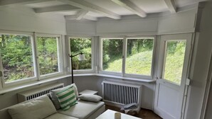 Une pièce avec un canapé blanc, une petite table et une chaise rose. De grandes fenêtres offrent une vue sur la verdure à l’extérieur.