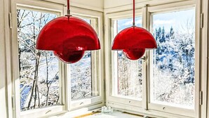 Un coin salon confortable avec un banc rembourré et une table en bois, de grandes fenêtres offrant une vue sur le paysage enneigé, et deux lampes suspendues rouges.