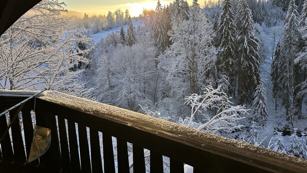 Une vue depuis un balcon doté d’une balustrade en bois, donnant sur une forêt enneigée et sur un soleil lointain.