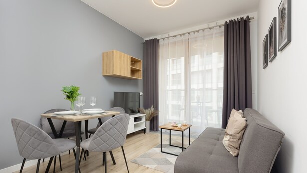 Una sala de estar moderna con un sofá gris, dos sillas grises, una mesa de comedor de madera con un jarrón de flores, una mesita auxiliar y un televisor.