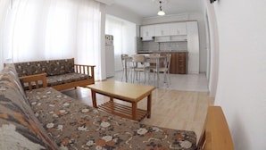 A living room with a sofa, a wooden table, and a kitchen area with cabinets and a dining table. - Minem Apart Otel (Fethiye)
