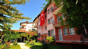 A multi-story building with balconies, a garden with red flowers, and a flag. - Minem Apart Otel (Fethiye)