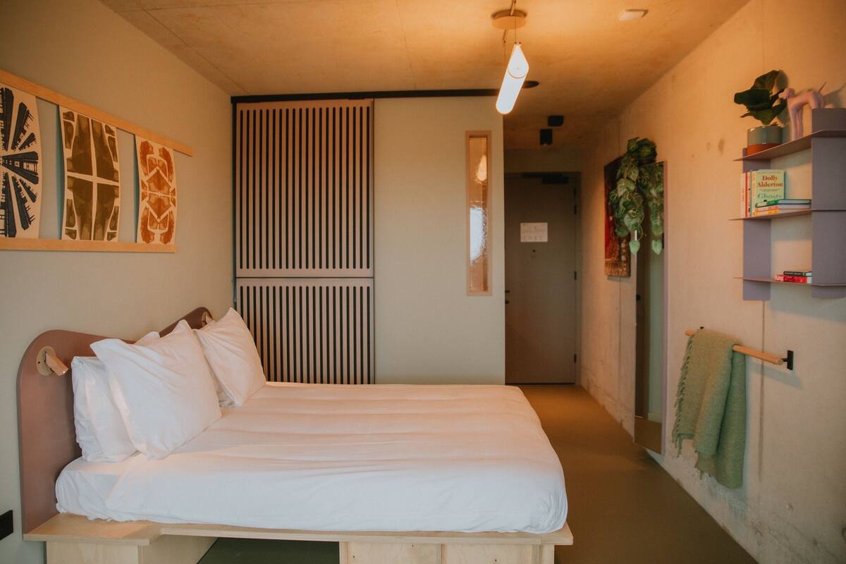 A bedroom with a bed, a radiator, a shelf with books, and a plant.