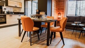Un espace repas moderne avec une table en bois et des chaises orange, une cuisine en arrière-plan et une vue sur l’extérieur grâce à de grandes fenêtres.