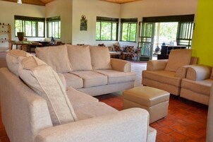 Amplia sala de estar con un sofá seccional color beige, un pequeño reposapiés y una mesa de comedor con sillas. Amplias ventanas ofrecen luz natural y vistas a zonas verdes exteriores.