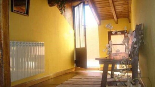 Une pièce aux murs jaunes, avec un plafond en bois, un radiateur, une fenêtre avec des stores et une table sur laquelle est posé un vase de fleurs.
