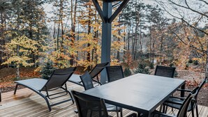Une terrasse en bois à l’extérieur, avec une table et des chaises, entourée d’arbres aux feuilles d’automne.