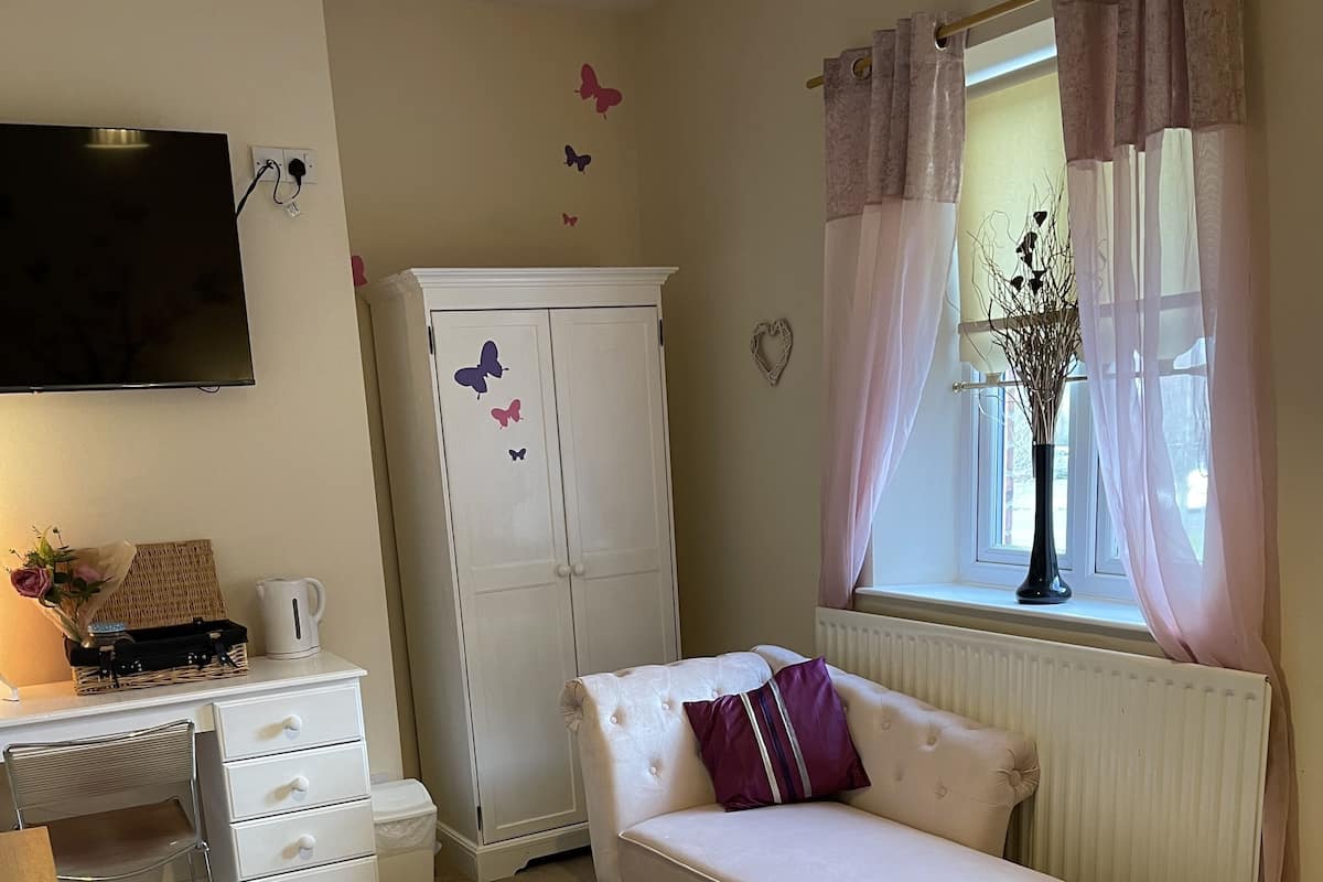 A bedroom with a bed, a sofa, a desk with drawers, a wardrobe, a window with curtains, and a flat-screen TV mounted on the wall.