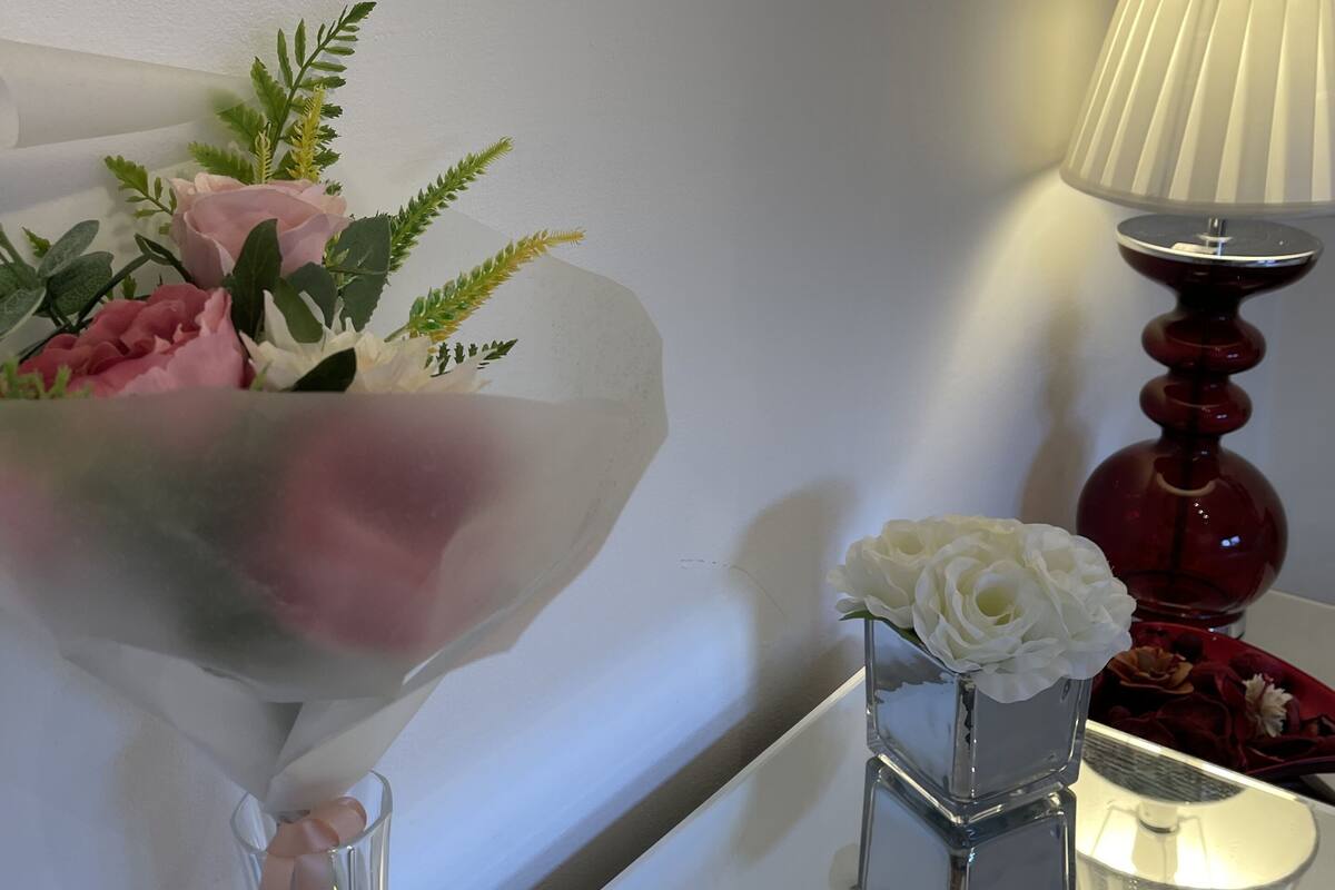 A bouquet of flowers in a glass vase, a small white vase with a single white rose, and a red lamp on a table.