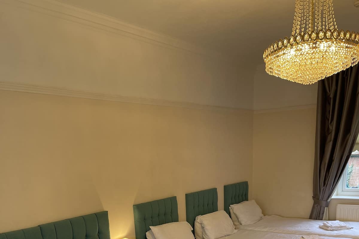 A hotel room with two beds, each with white linens and pillows, a green upholstered headboard, a chandelier, and a window with curtains.