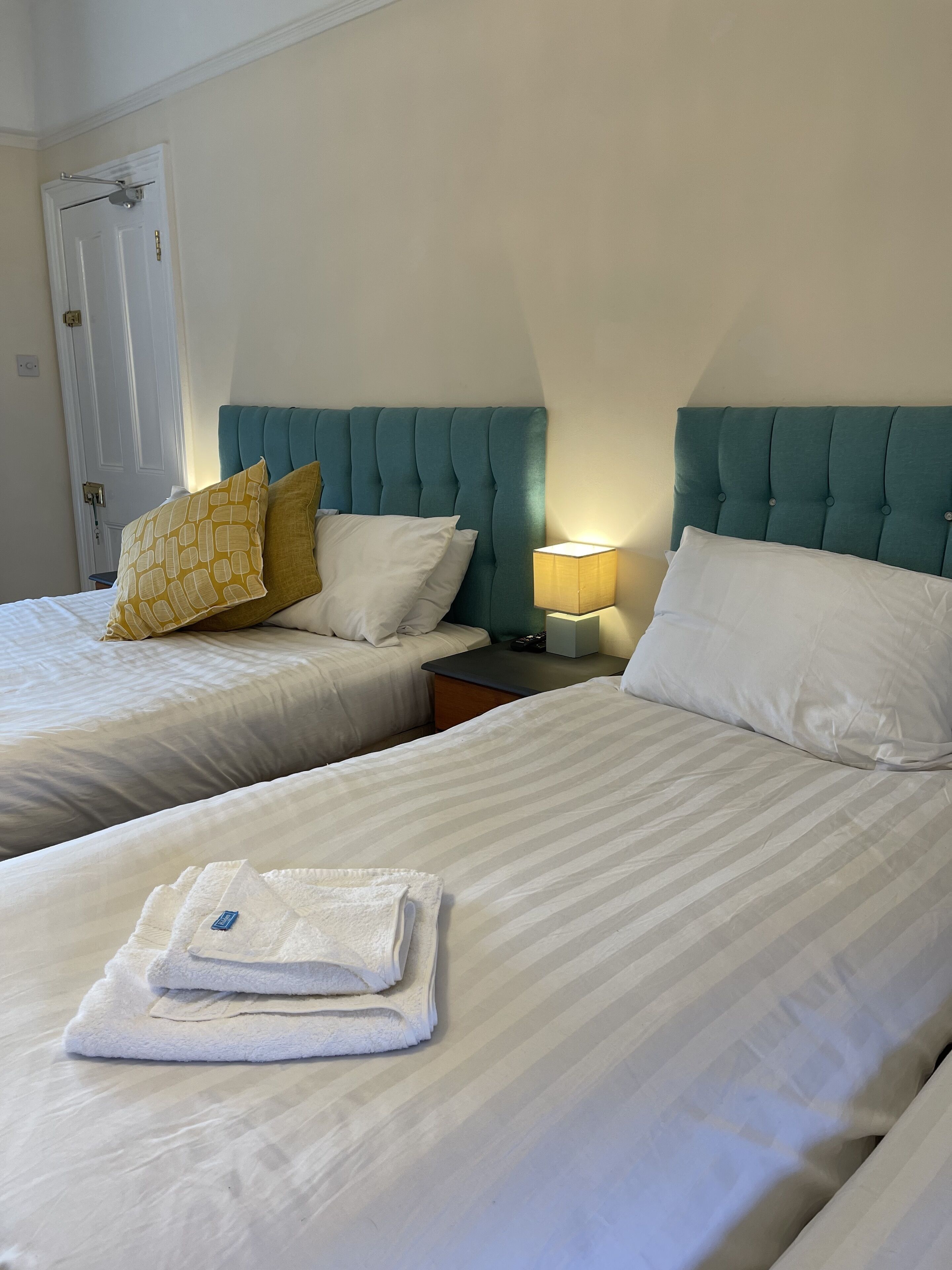 A hotel room with two beds, each with white bedding and a teal headboard. There is a folded towel on the bed in the foreground.