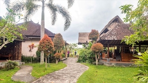 A pathway leading to traditional-style buildings with thatched roofs, surrounded by lush greenery and palm trees. - AlamGangga Villas Tirta Gangga (Karangasem)