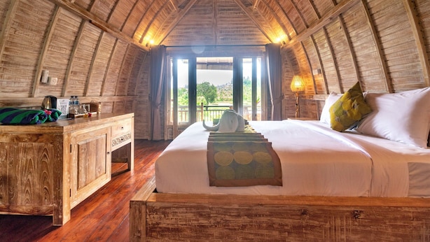 A cozy, wooden-paneled room with a bed, bedside table, and a view through a large window. - AlamGangga Villas Tirta Gangga (Karangasem)