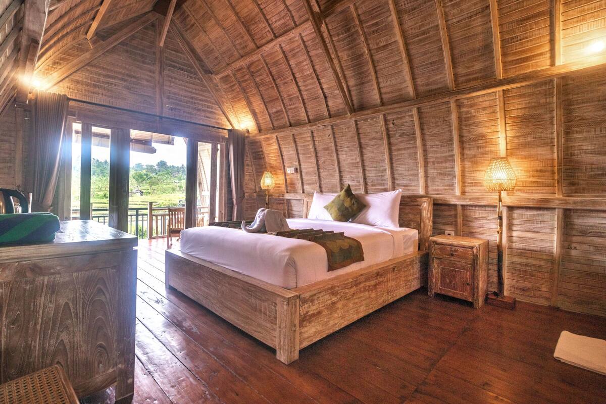A rustic wooden room with a large bed, a desk, and a view of the outdoors.