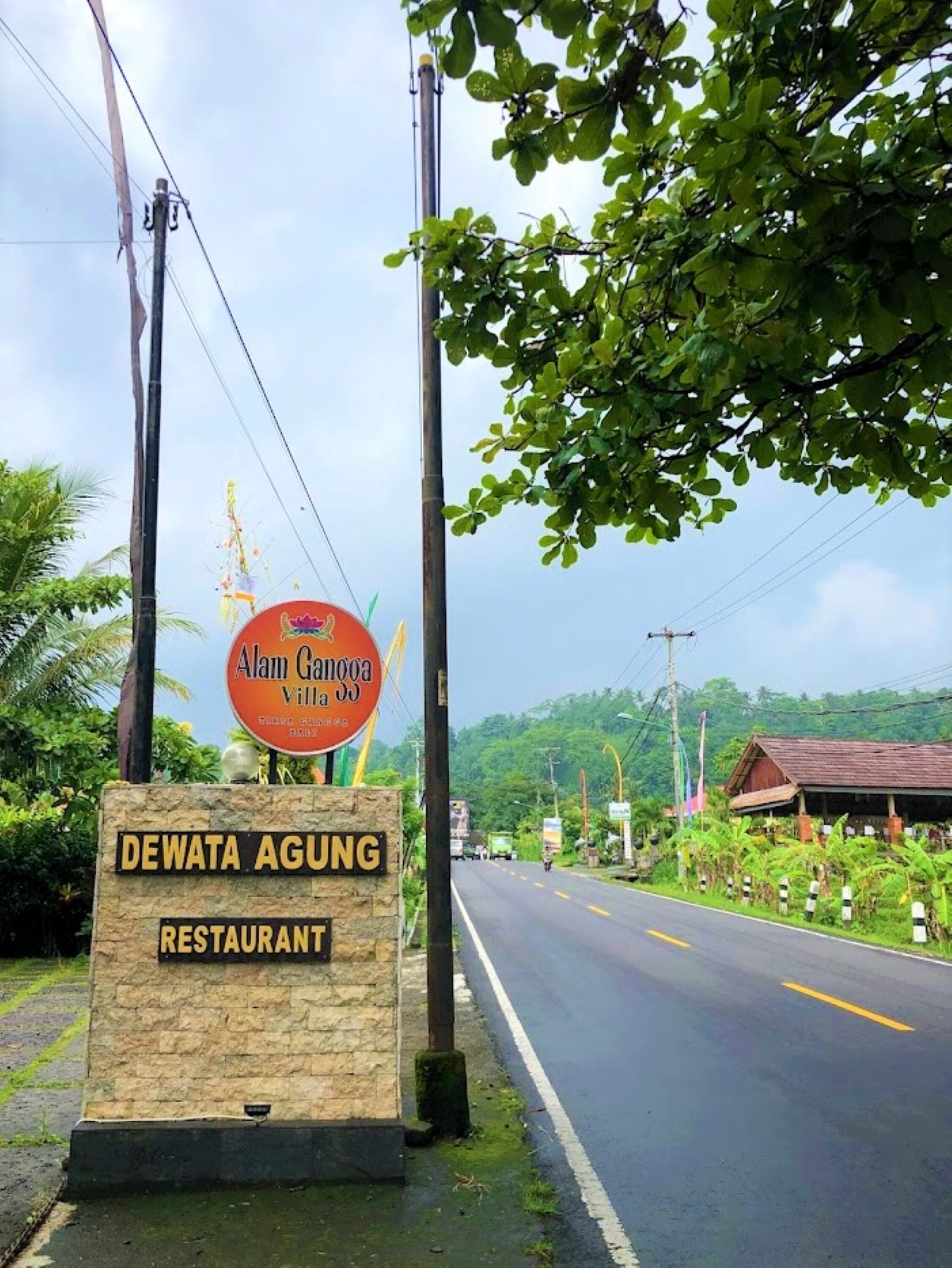 A sign for Dewata Agung Restaurant with a view of a road and greenery in the background.