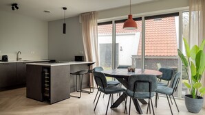 Een moderne keuken met een centraal eiland, een eethoek met een ronde tafel en stoelen, en grote ramen met uitzicht op een dak met rode tegels.