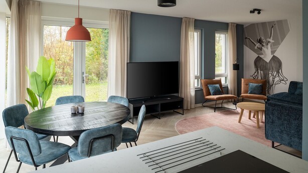 Een moderne woonkamer met een ronde eettafel, blauwe stoelen, een flatscreen televisie en een groot raam met gordijnen.