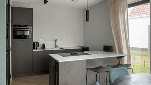 Een moderne keuken met een inbouwoven, een spoelbak en een klein eiland met waterkoker en broodrooster. Op de voorgrond staan twee stoelen en een tafel.