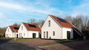 Een rij witte huizen met rode daken en zonnepanelen, gelegen in een woonwijk met een verharde weg en bomen op de achtergrond.