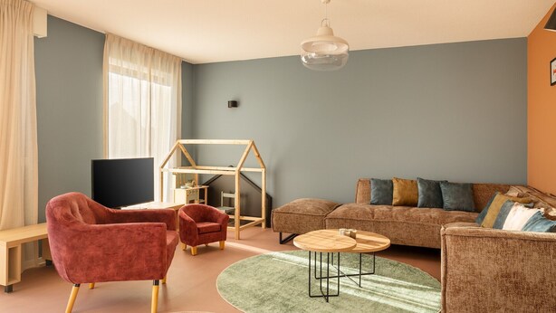 Uma sala de estar com sofá, duas poltronas vermelhas, uma mesa de madeira, um tapete verde e uma casinha de brincar.