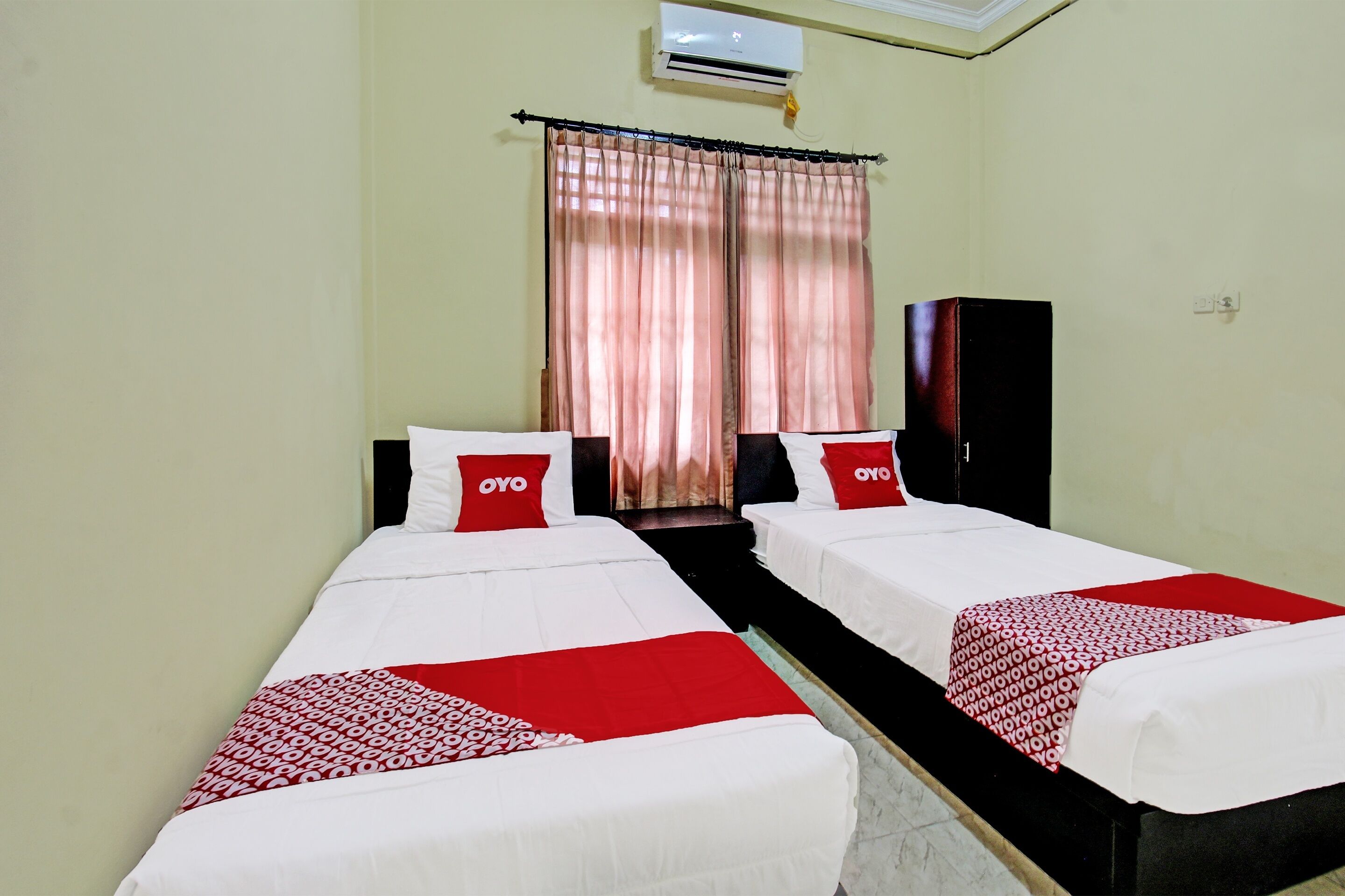 Habitación de hotel con dos camas, cada una con cubrecama y almohadas rojas y blancas, un armario negro y una ventana con cortinas rosas.