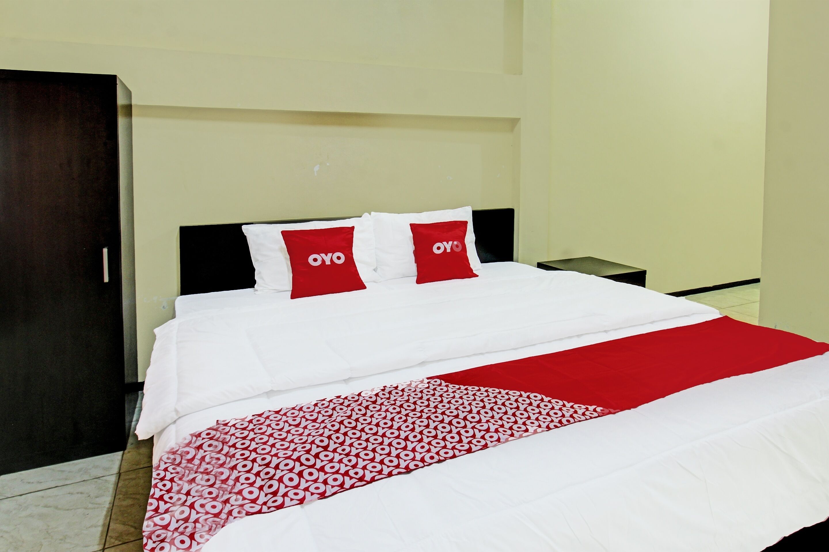 Une chambre d’hôtel comprenant un lit, deux oreillers à l’effigie de la marque « OYO », une table de chevet et une armoire.