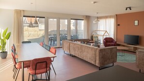 A modern living room with a sofa, dining table, chairs, a TV, and a large window with a view of the outside. - Child-friendly 10-bed holiday home in Kids Quarter (Nieuwvliet-Bad)