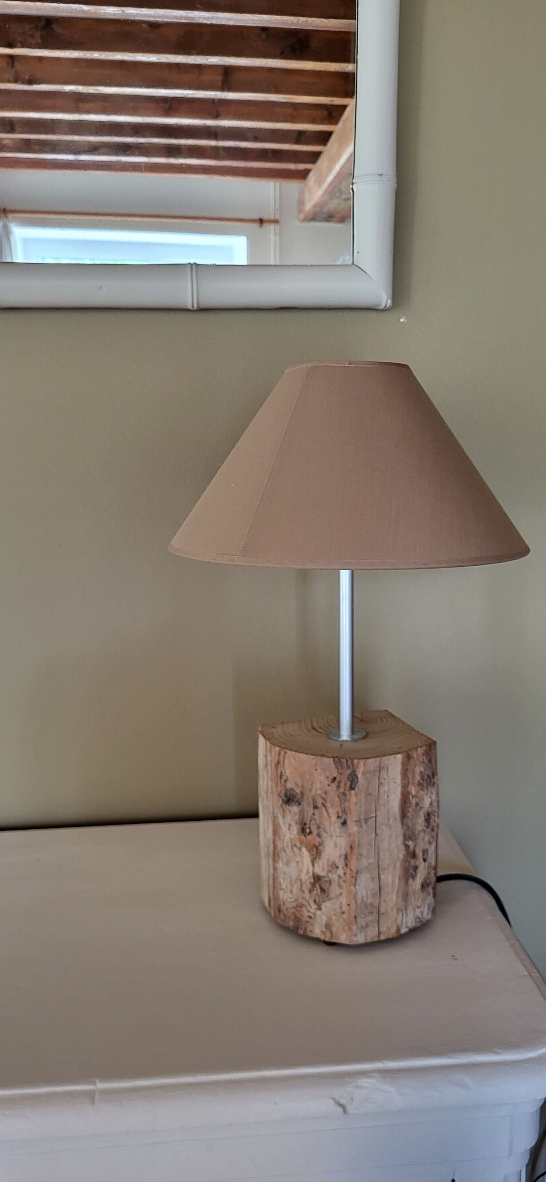 A lamp with a wooden base on a white surface, a window with blinds above, and a plain wall.