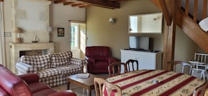 Un salon avec une cheminée, un canapé à carreaux, un fauteuil rouge, une table à manger recouverte d’une nappe et un téléviseur posé sur un meuble.