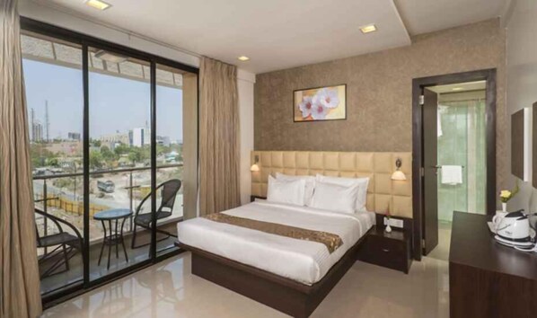 Quarto de hotel com uma cama grande, uma cadeira, uma escrivaninha e vista da cidade através de amplas janelas.