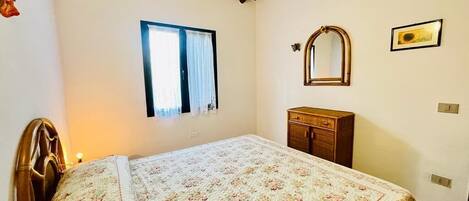 A bedroom with a bed, a window, a wooden cabinet, and a mirror on the wall.