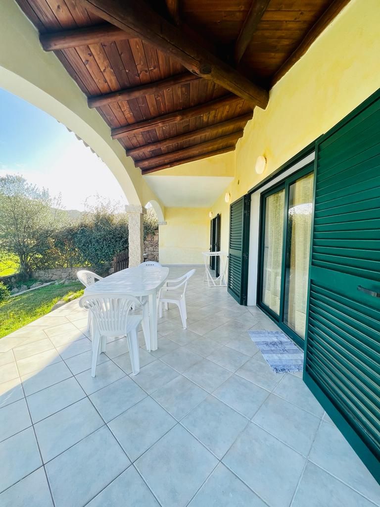 Um pátio externo com mesa e cadeiras, área coberta com teto de madeira e uma porta com persianas verdes.