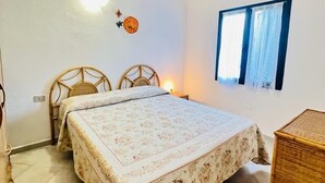 A bedroom with a bed, two chairs, a wooden cabinet, a window with curtains, and a wall-mounted light. - Casa Vacanza L'alzoni (San Teodoro)