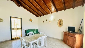 A dining area with a wooden ceiling, a table with chairs, a green sofa, a wooden cabinet with a television, and decorative wall items. - Casa Vacanza L'alzoni (San Teodoro)