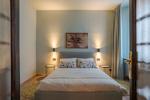 Ein Schlafzimmer mit einem Bett, zwei Nachttischlampen, einem Wandbild und Blick durch eine offene Tür.